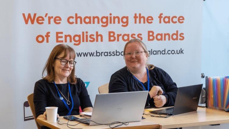 Two women with brown hair and wearing black tops with blue lanyards siting in front of a sign that read's "We're changing the face of English Brass Bands"