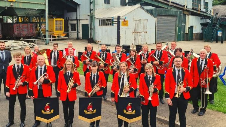Hade Edge band stand in front of industrial buildings at the Coal Museum site. They are wearing red jackets and holding their instruments