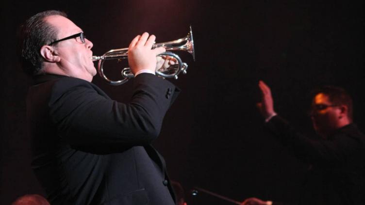 Side-profile of paul playing a soprano cornet. He is wearing a black jacket and glasses. The room is dark and a conductor can be seen in the distance