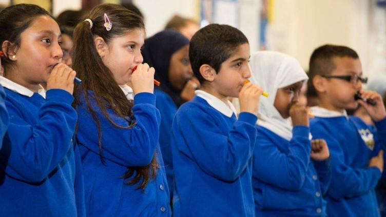 Five children holding different coloured plastic mouthpieces to their mouths. They are wearing blue sweatshirts and white colors