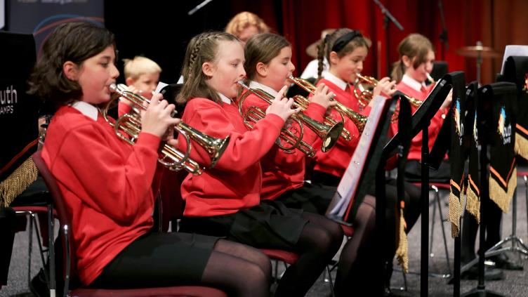 Line of girls playing brass insturments, they are wearing red sweatshirts and black trousers or skirts