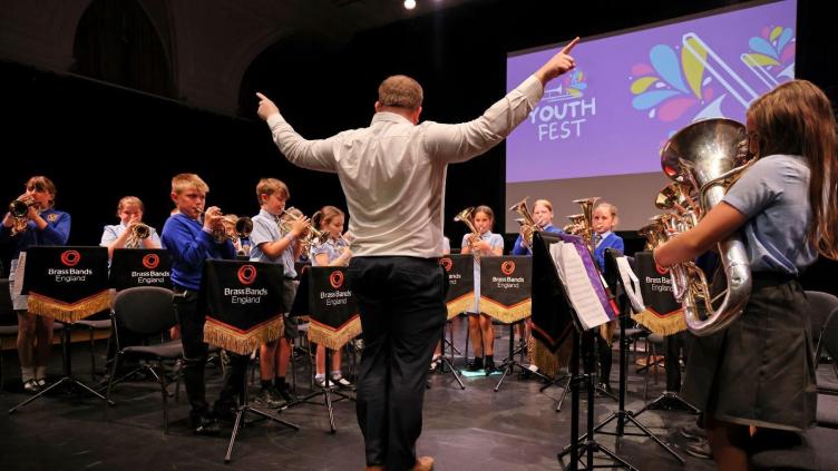 A children's brass band being conducted playing instruments while standing in front of a Youth Fest banner