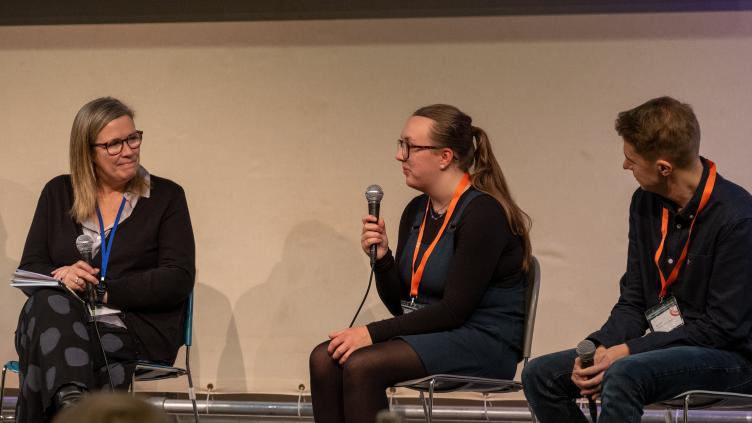 Two women and a man sit on stage, one holding a microphone speaking