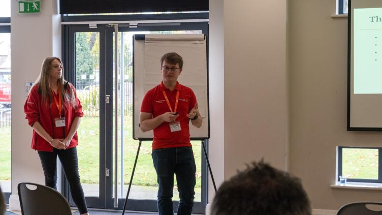 Two people in red jackets stand in front of a screen