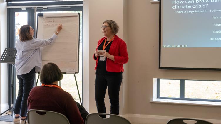 Woman in red jacket presents to audience in front of screen, woman in blue shirt writes notes on paper 