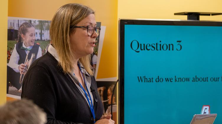 Blonde haired woman in glasses presents in front of screen with blue background