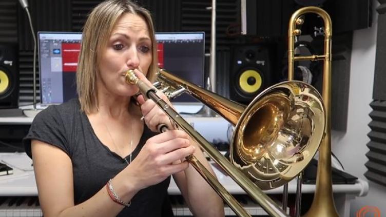 Carol has short blond hair and is earing a grey t-shirt. She is standing in front of of a computer screen and speaker and is playing the trombone