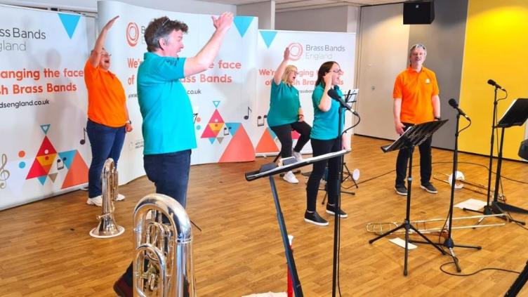 Five musicians each holding a brass instrument and wearing a different brightly coloured t-shirt are waving their arms in the air