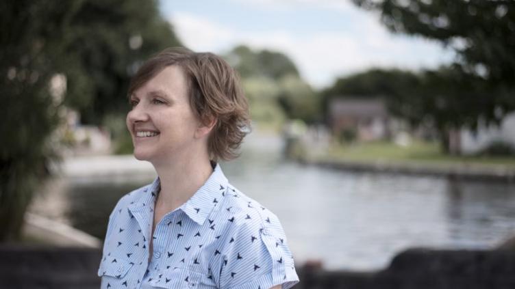 Liz is standing in front of a lake and looking to the left. She has short brown hair and is wearing a blue shirt