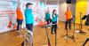 Five musicians each holding a brass instrument and wearing a different brightly coloured t-shirt are waving their arms in the air